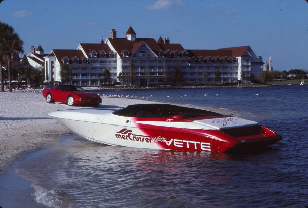 A red corvette is parked on a beach at the water's edge next to a red and white speed boat. In the background is a hotel resort.