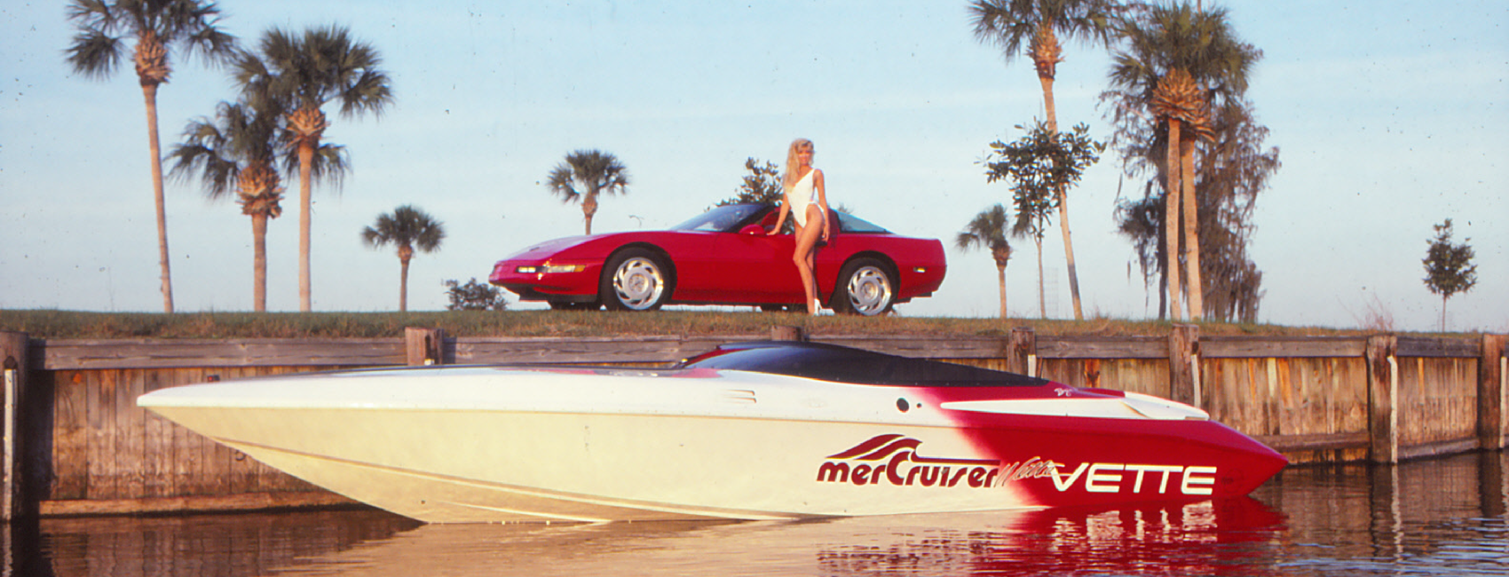 A model stands next to a red Corvette surrounded by palm trees. In the foreground a red and white speed boat is floating in the water.